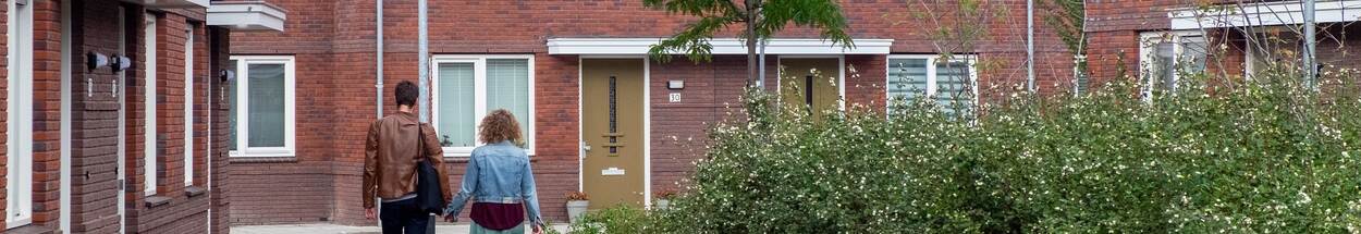 Een jong stel loopt hand in hand in een straat met nieuwbouwhuizen en een groen plantsoen.