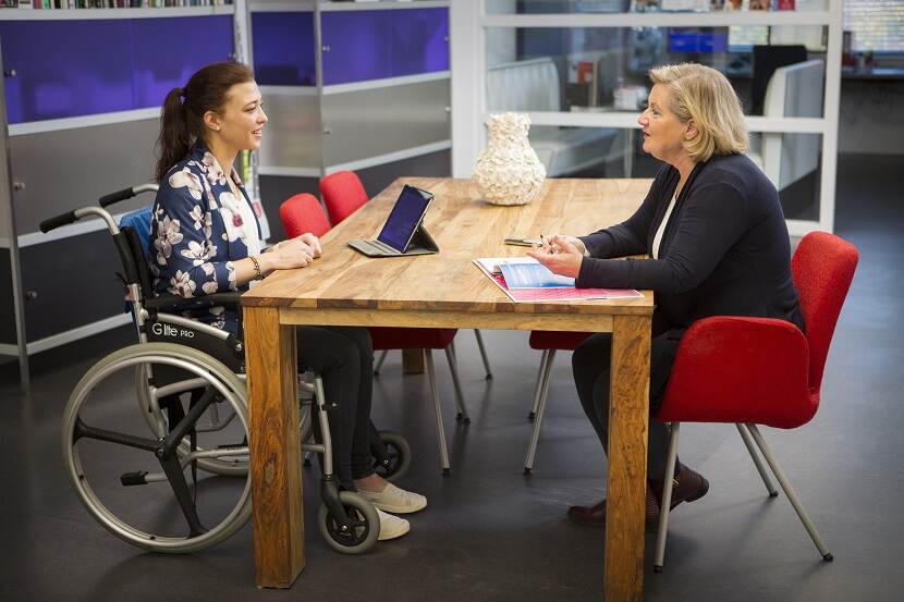 Twee vrouwen zitten aan tafel en voeren een gesprek. De vrouw links zit in een rolstoel.
